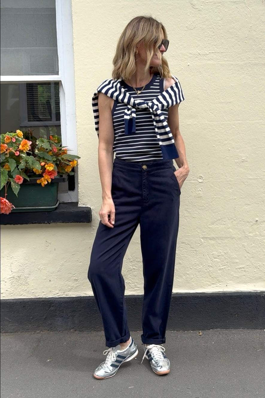 Woman wearing a striped shirt and navy trousers standing against a light-colored wall.