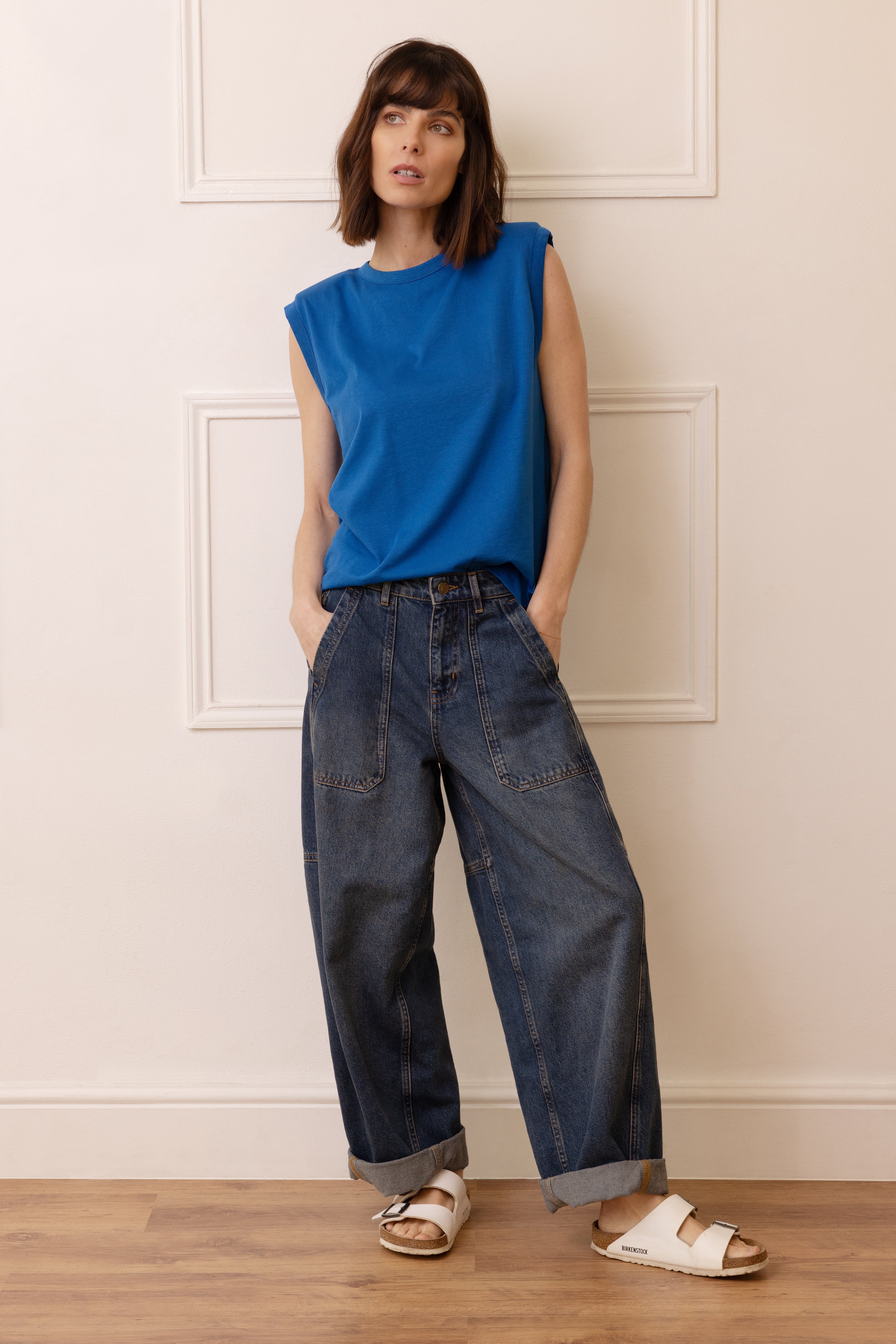 Woman wearing a blue sleeveless top and dark jeans standing against a white wall.