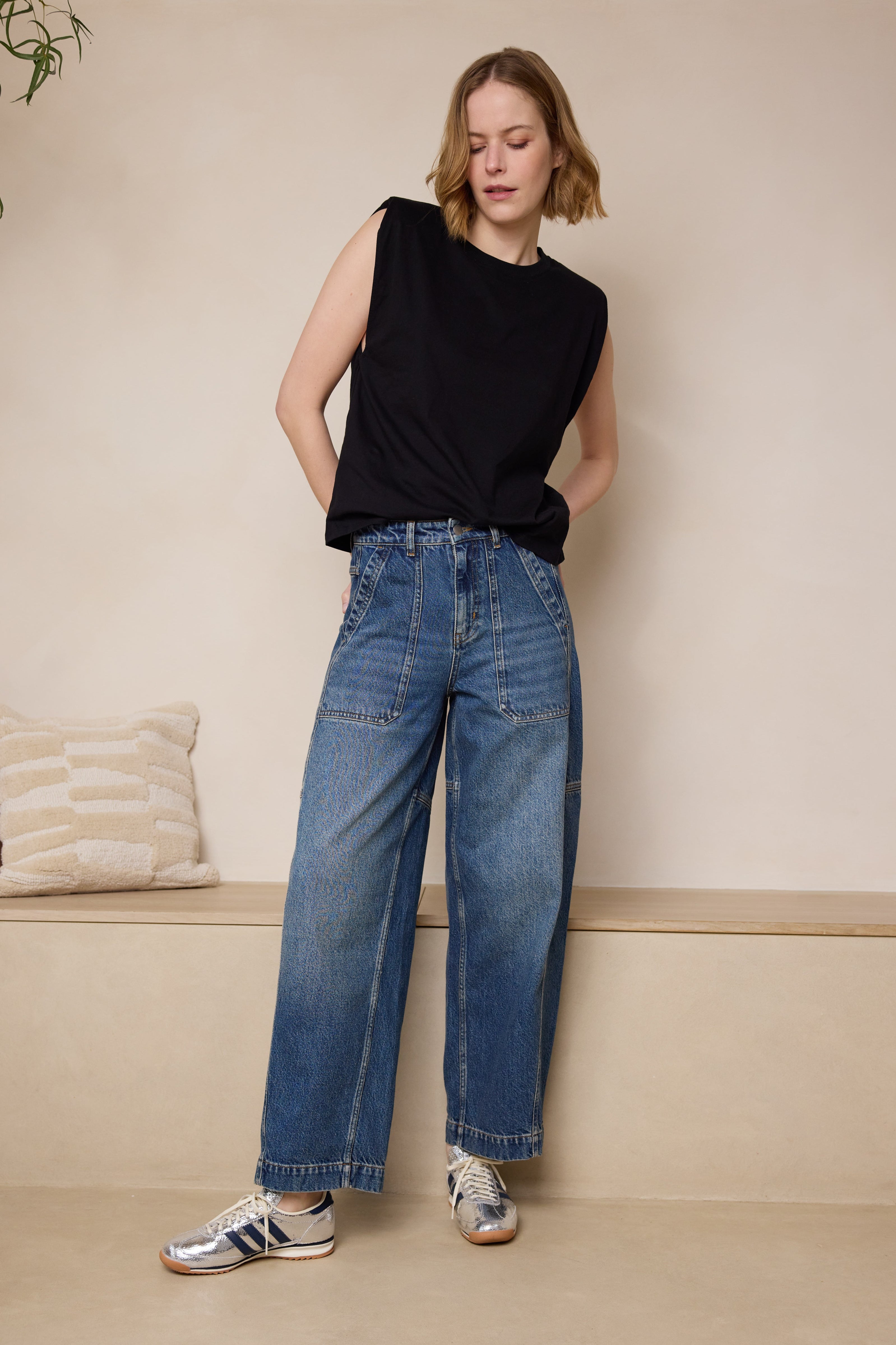 A woman standing in a room wearing a black sleeveless top and blue barrel leg jeans with a rollable hem.