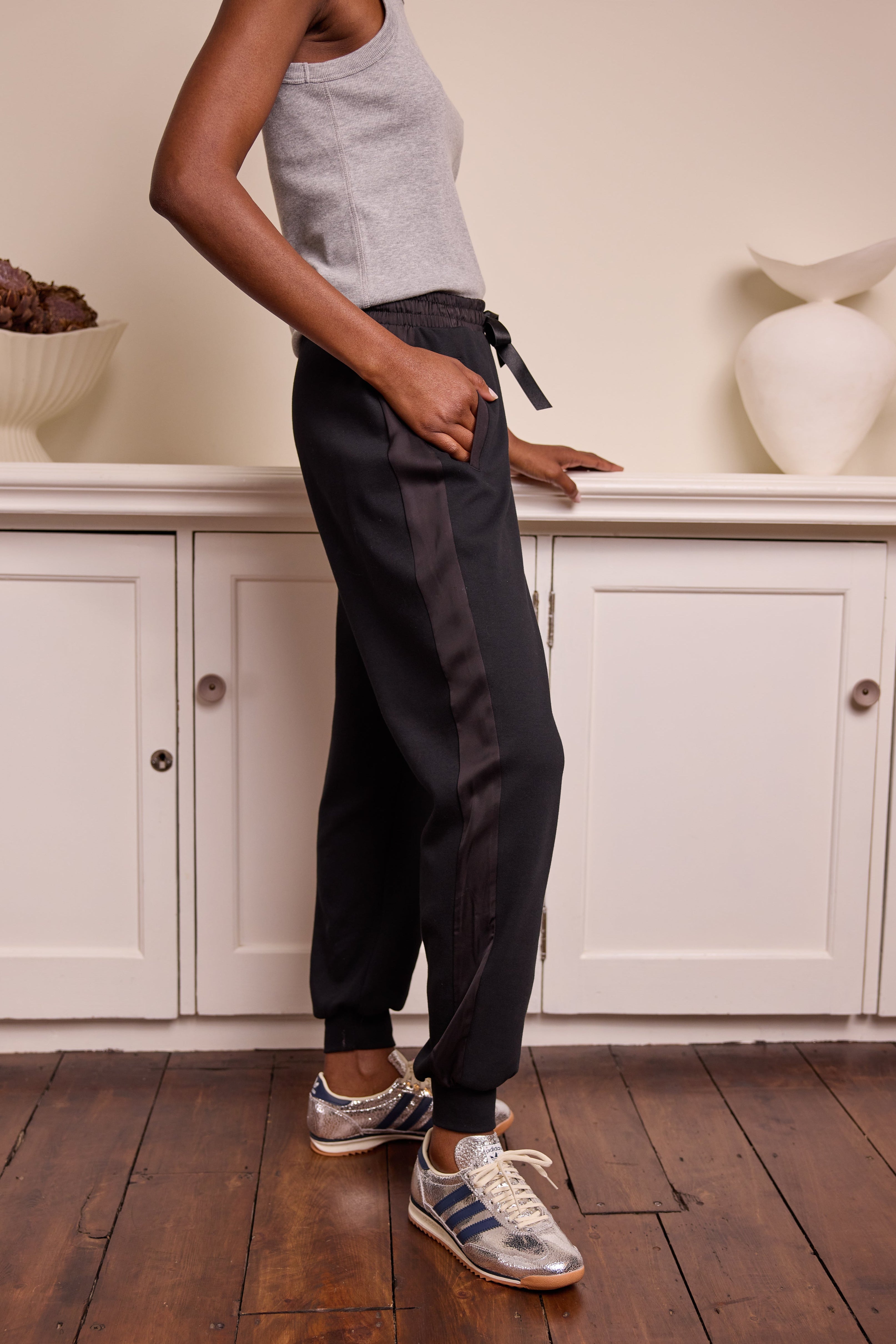 A person standing side-on wearing black side stripe joggers with a satin side stripe detail, paired with a white tank top and white sneakers.