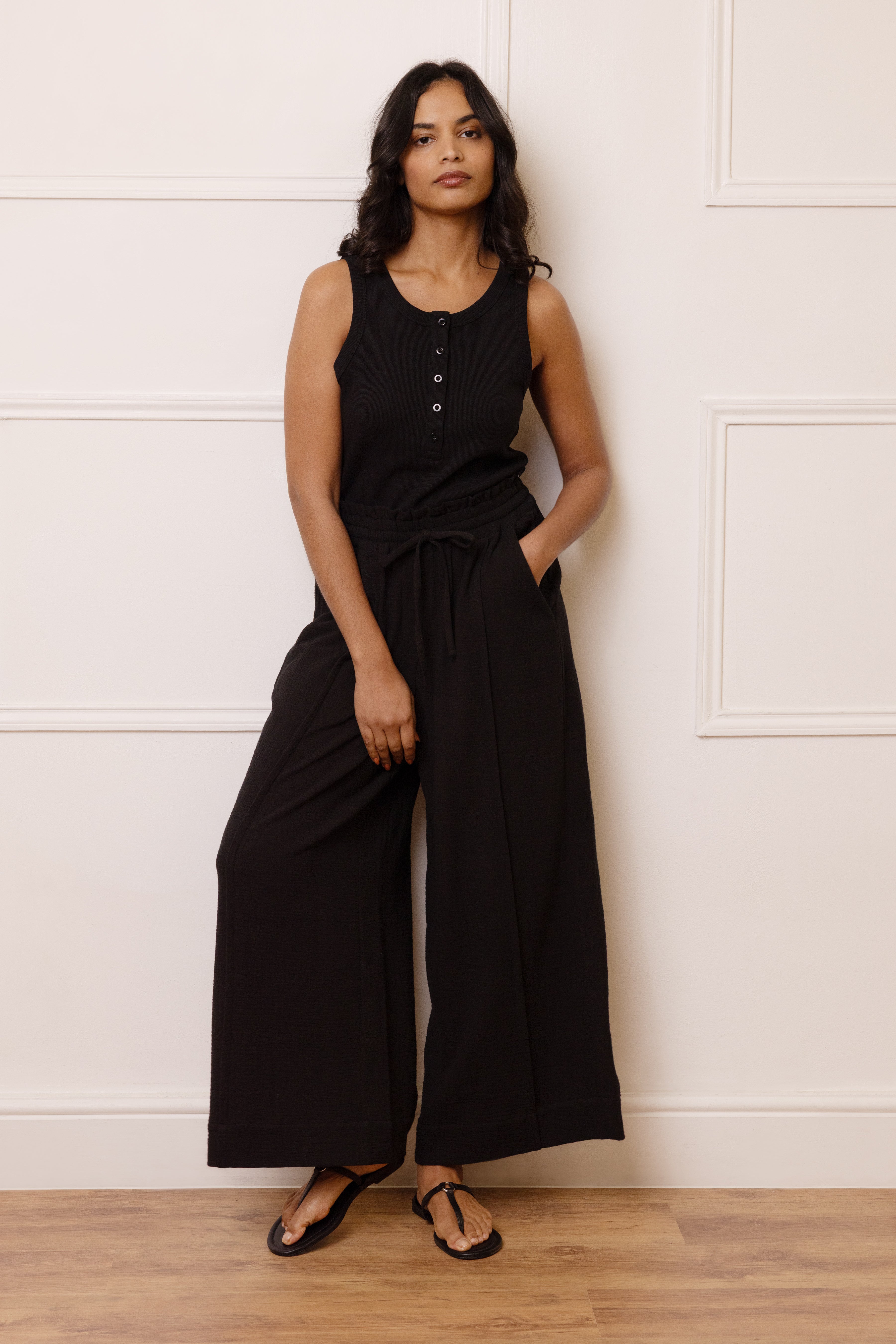 Woman wearing a black sleeveless jumpsuit against a white wall.