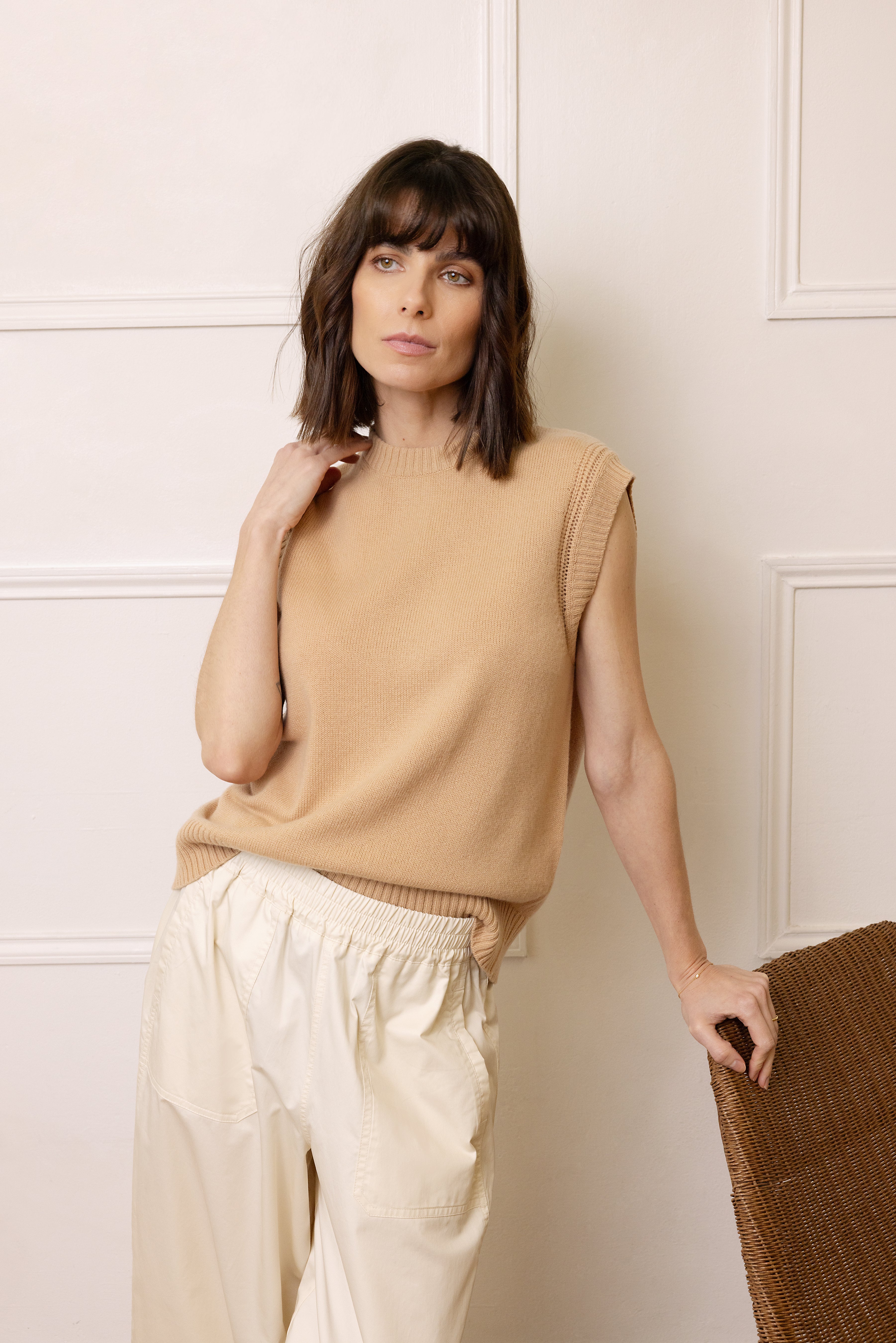 Woman wearing a beige sleeveless sweater and cream pants against a white wall.