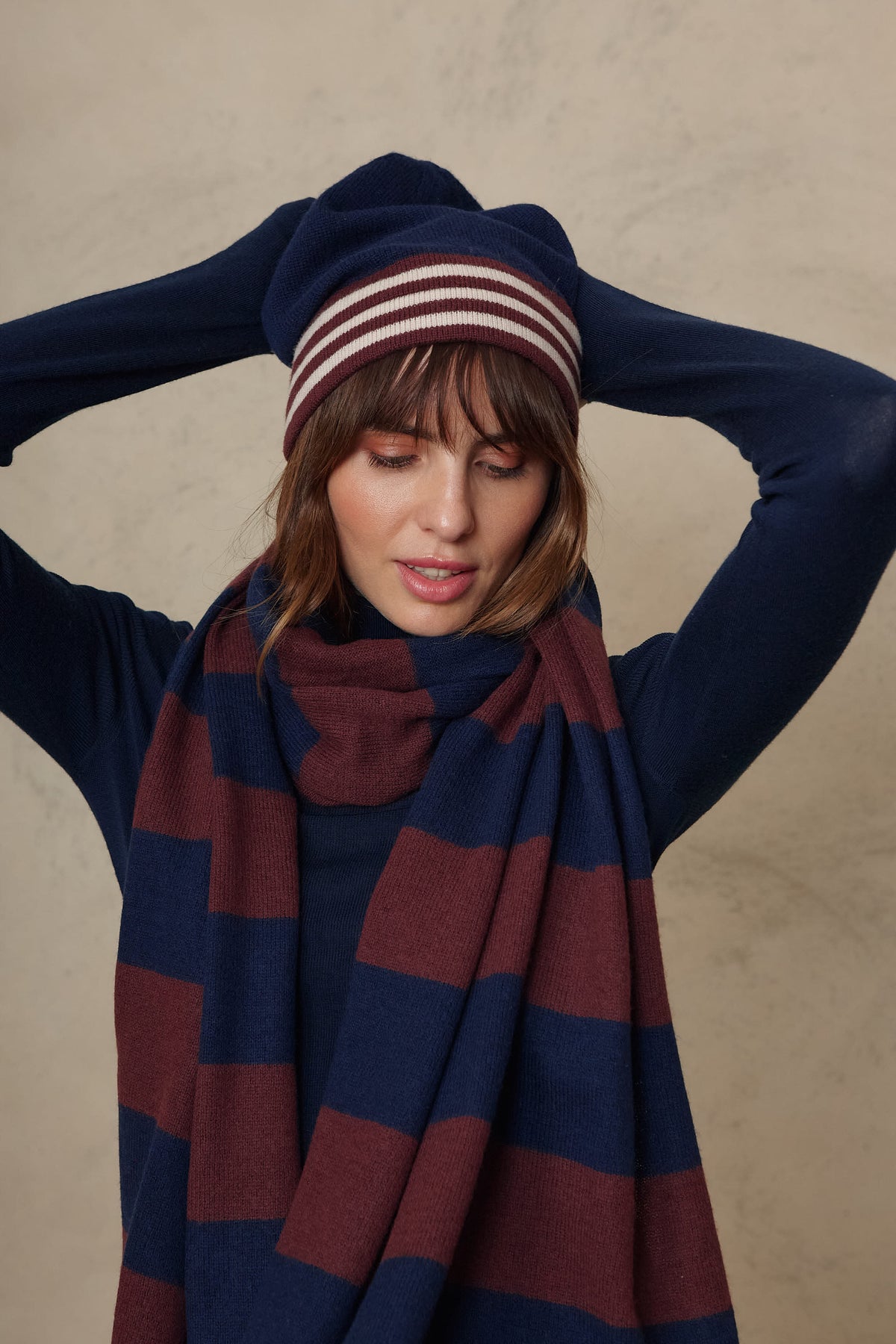 Model wearing the Carmel cashmere blend scarf in navy and burgundy with the Zinnie beanie in navy, hands behind her head