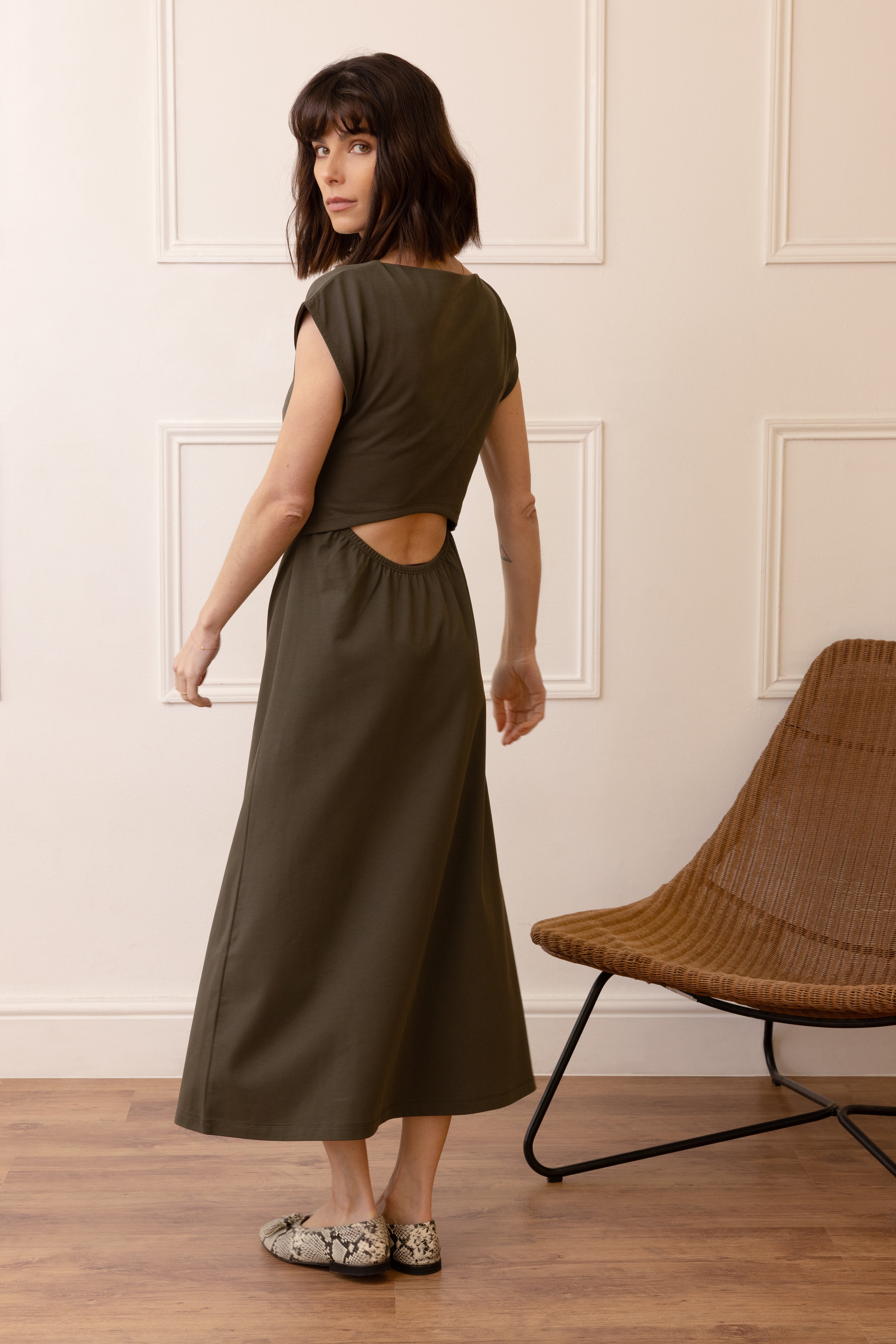 Woman wearing a green dress with an open back, standing in a room with wooden flooring and a wicker chair.