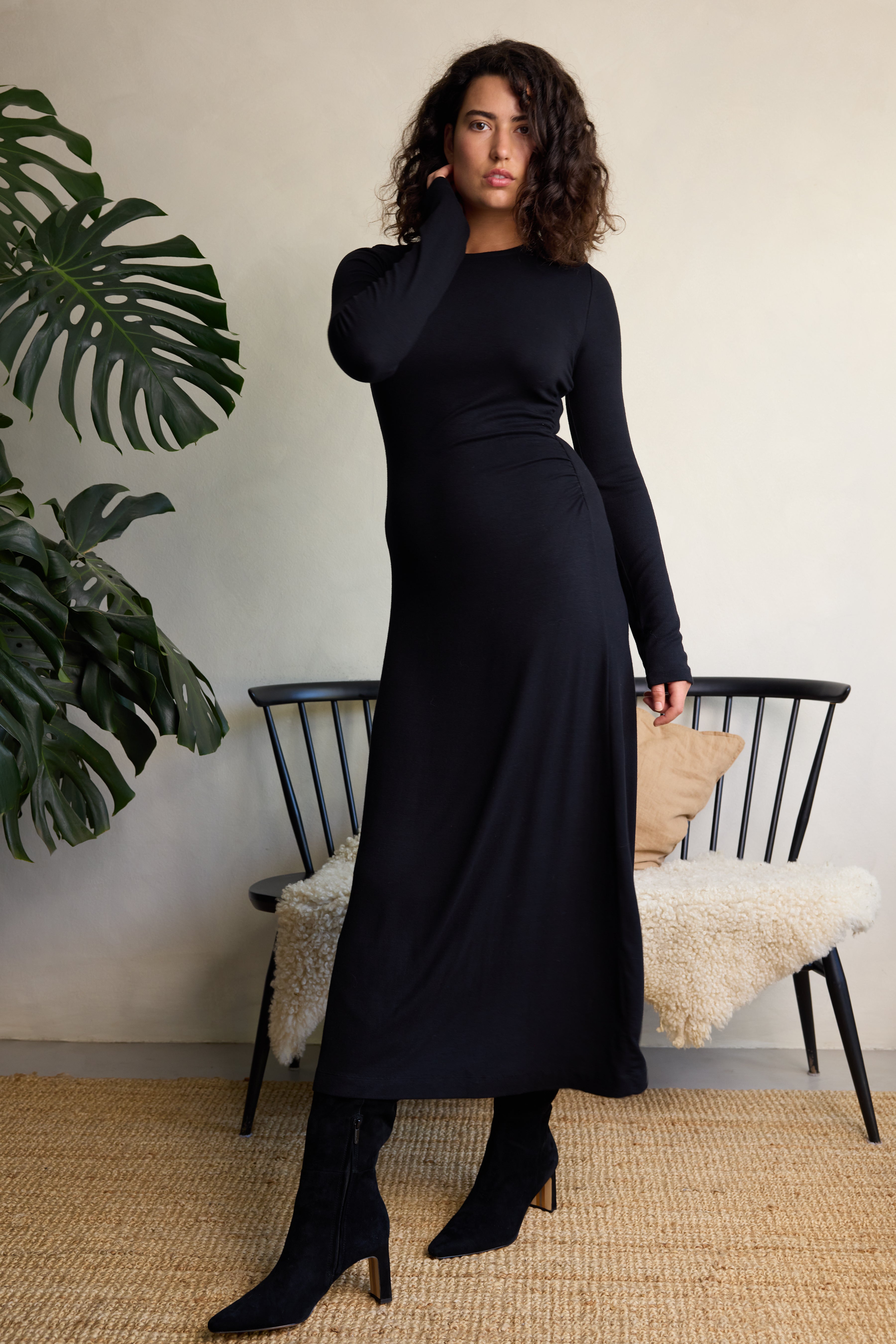 Woman wearing a black dress standing in a room with a plant and chair.