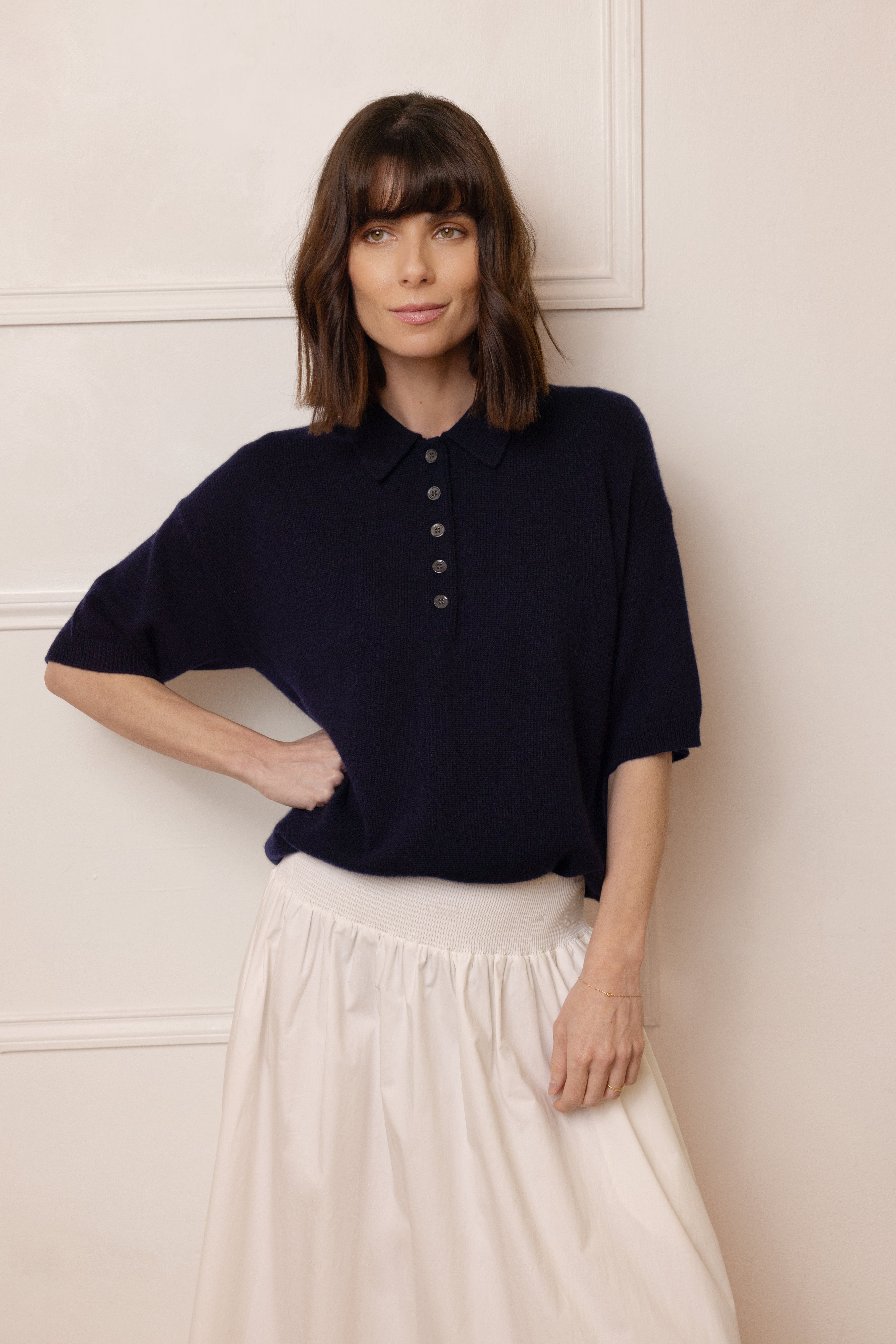 Woman wearing a navy blue polo shirt and white pants against a light-colored wall.
