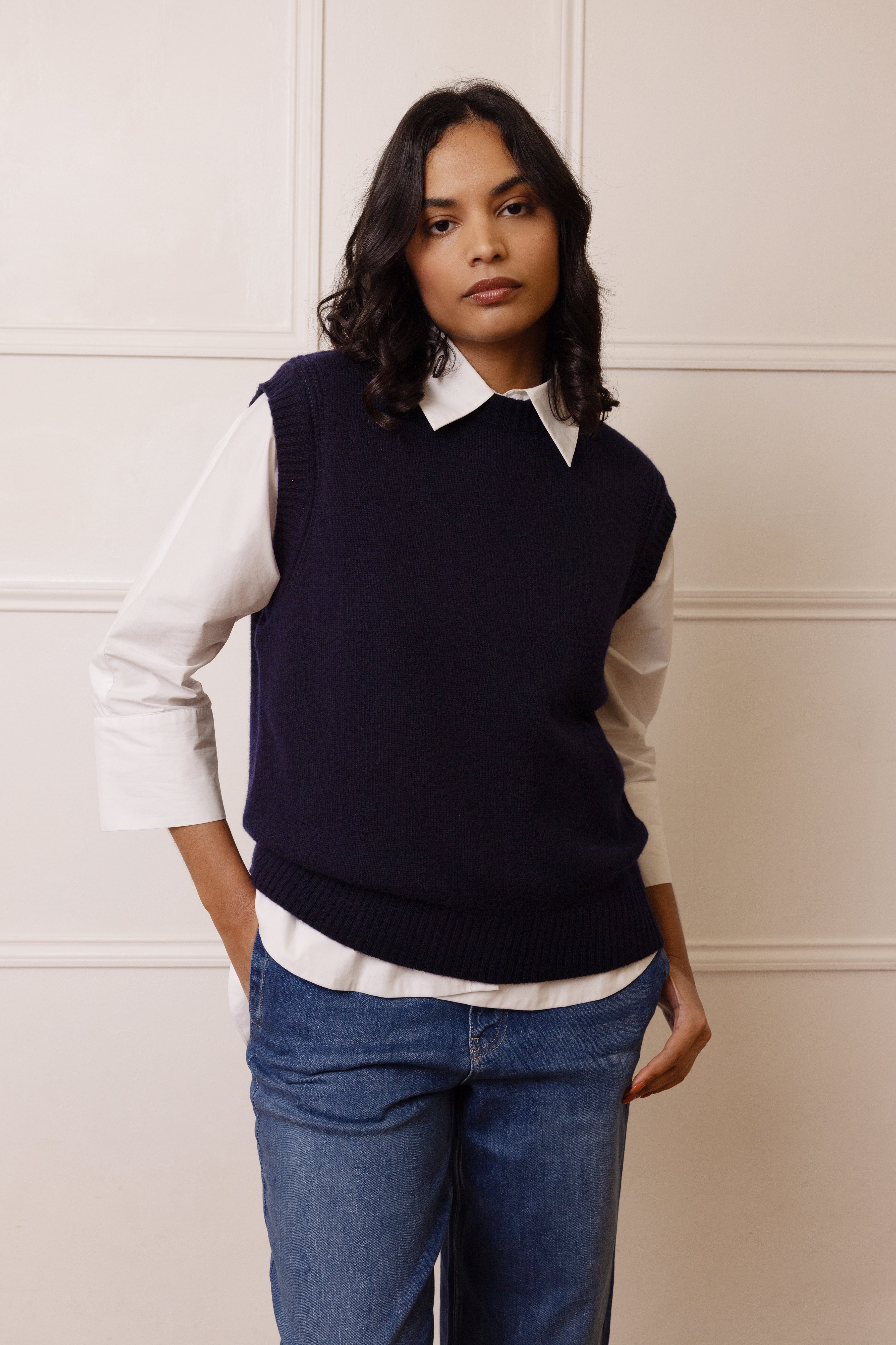 Woman wearing a navy sweater vest over a white shirt with blue jeans against a neutral wall.