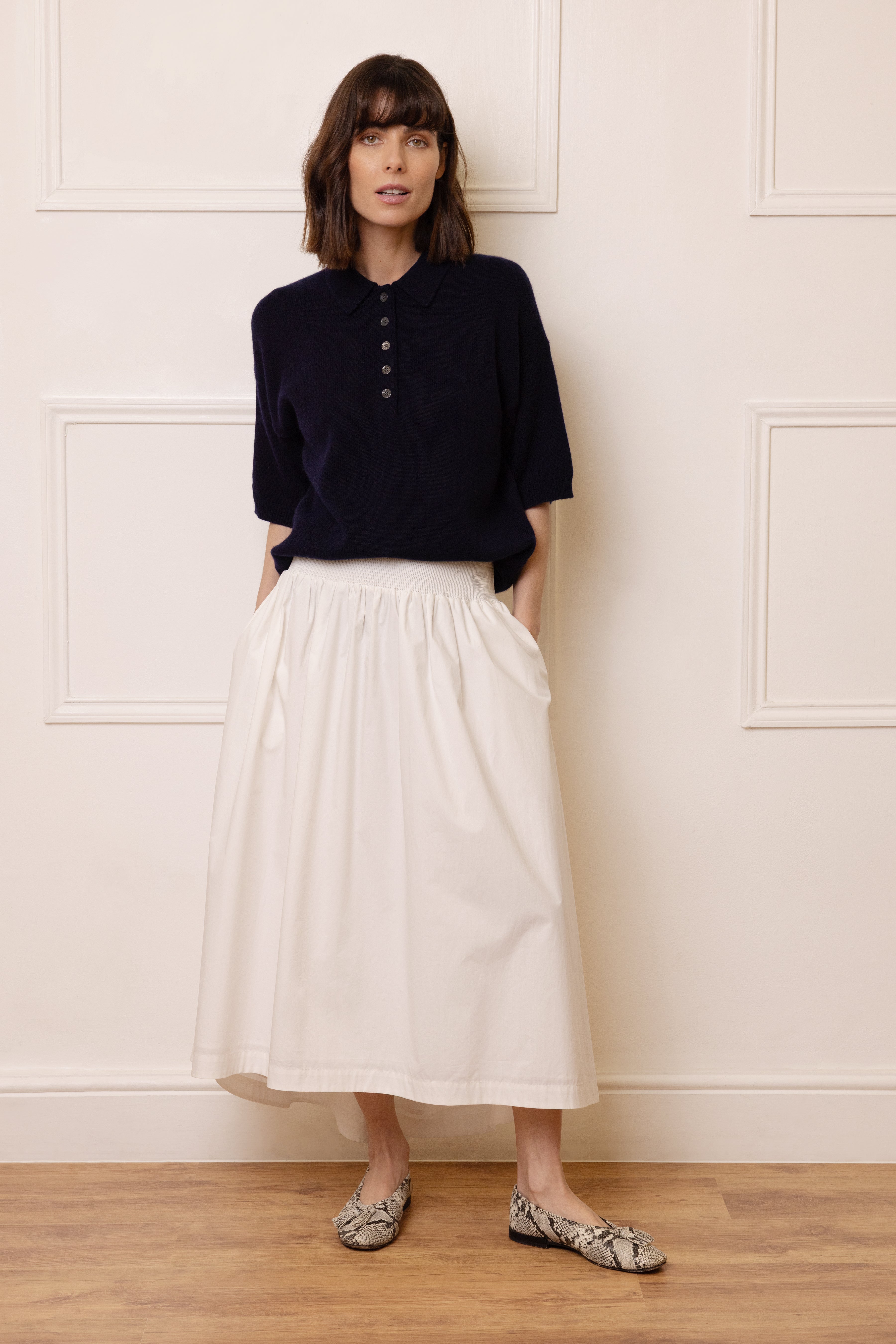 Woman wearing a navy top and white skirt against a white paneled wall.