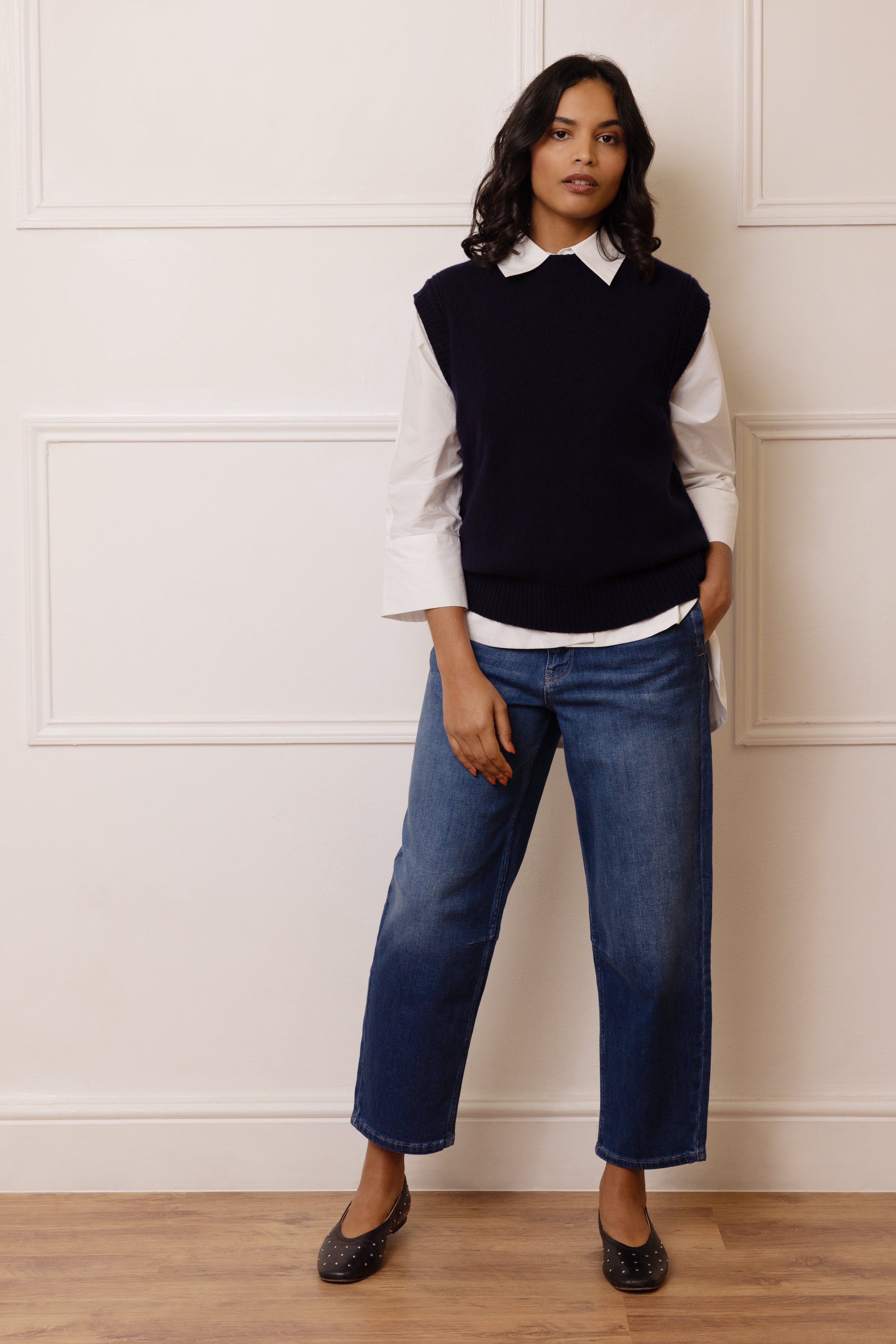 Woman wearing a navy sleeveless sweater over a white shirt with blue jeans against a white paneled wall.