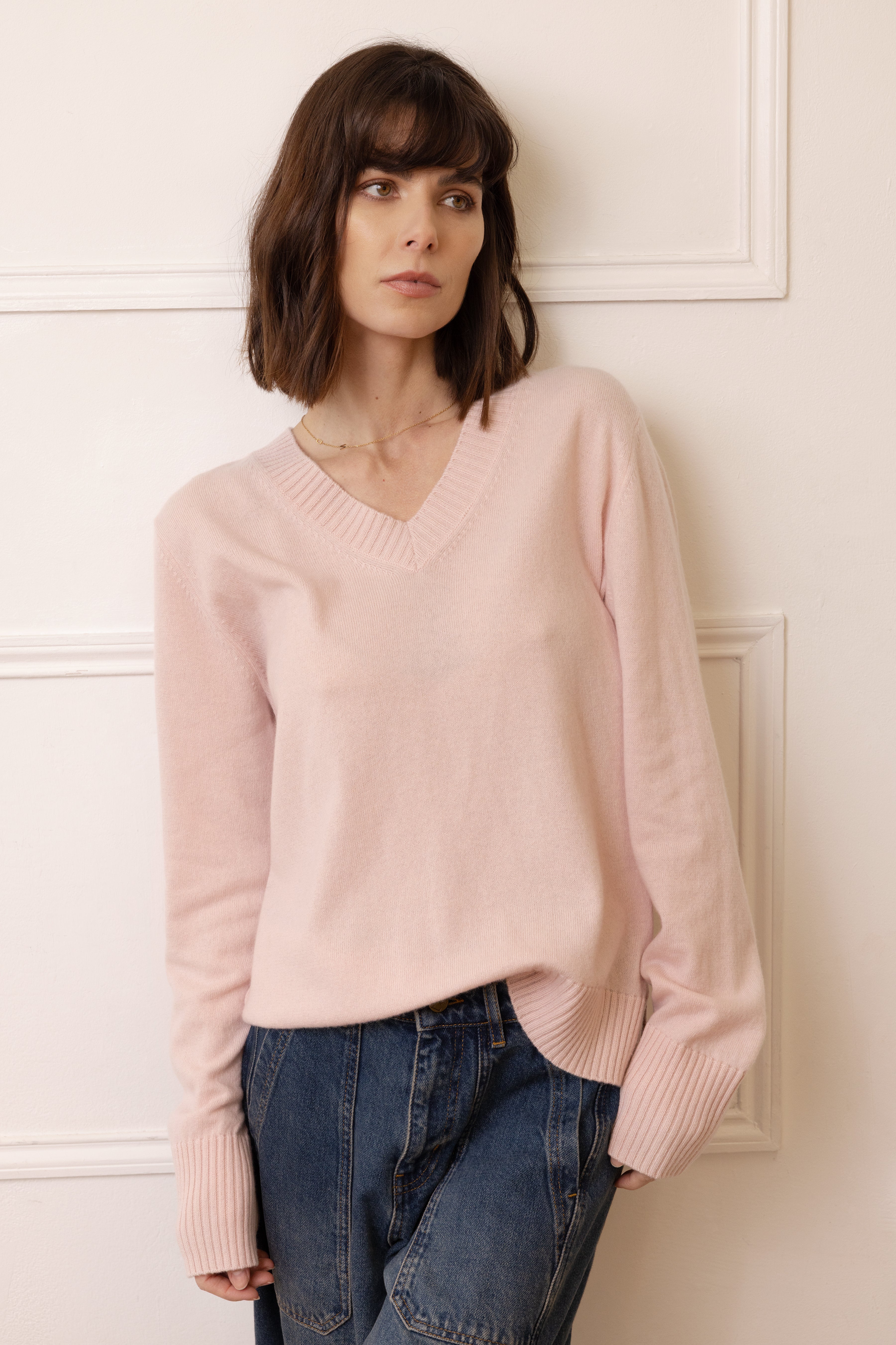 Woman wearing a light pink sweater and blue jeans against a white paneled wall.