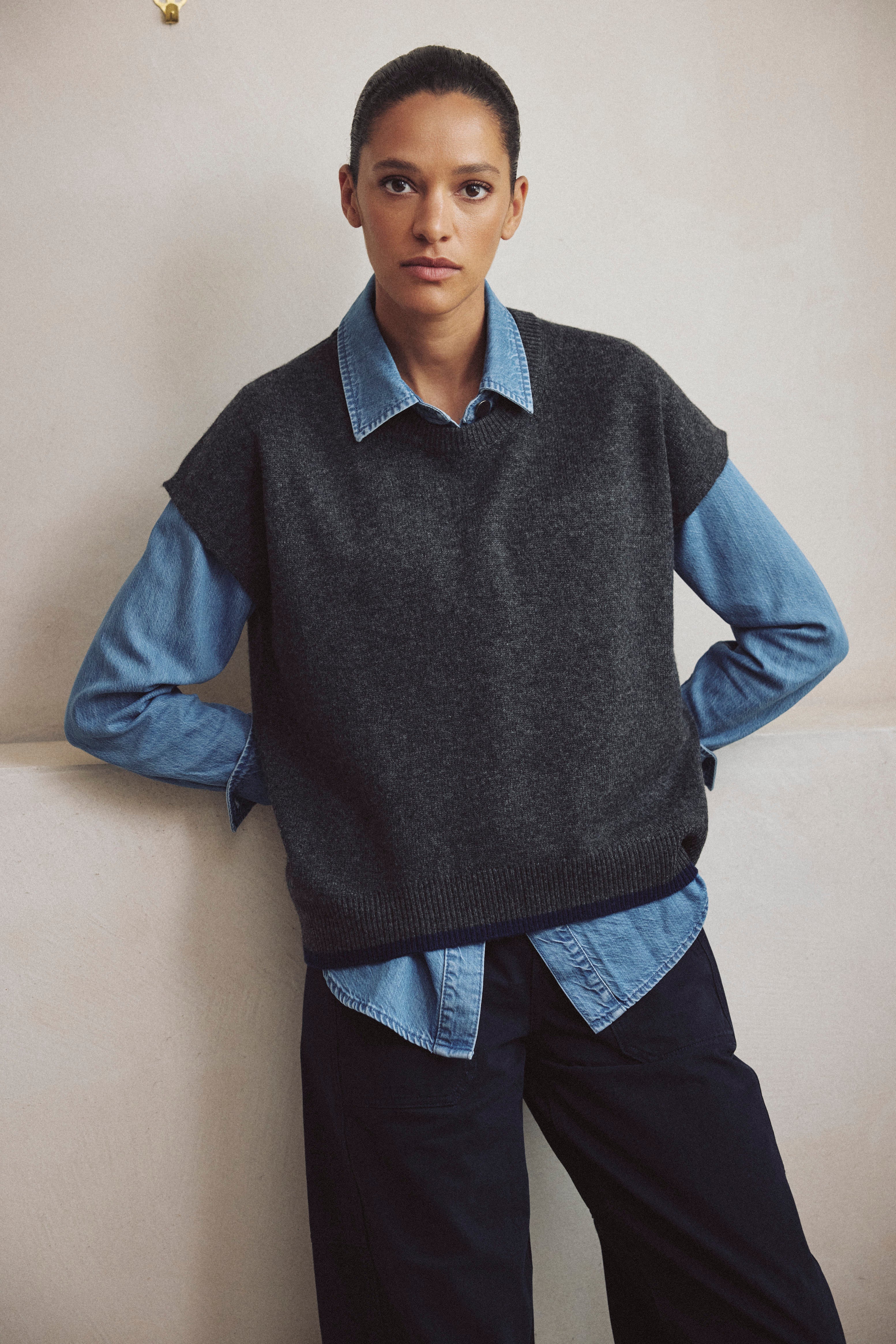 Ora charcoal knitted vest layered over a blue shirt with dark trousers