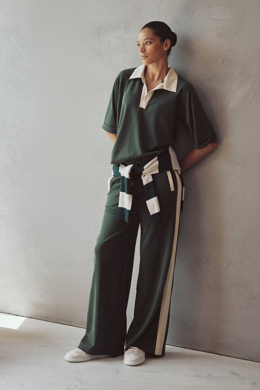 A woman stands against a textured gray wall wearing a dark green short-sleeve collared top with a cream collar, matching dark green wide-leg pants with cream side stripes and a matching tie belt, and white sneakers.