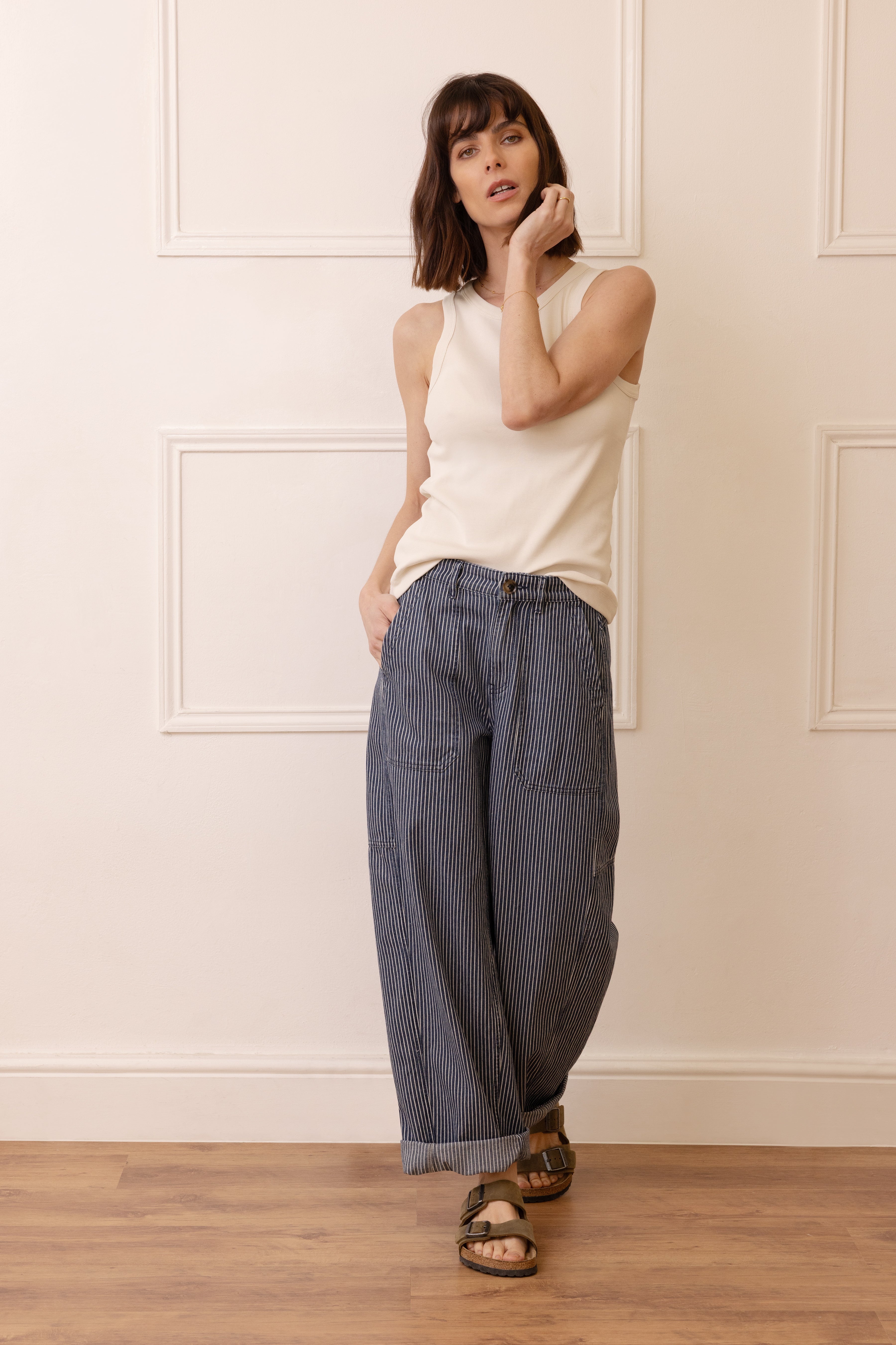 Woman wearing a white sleeveless top and blue striped pants against a white wall.