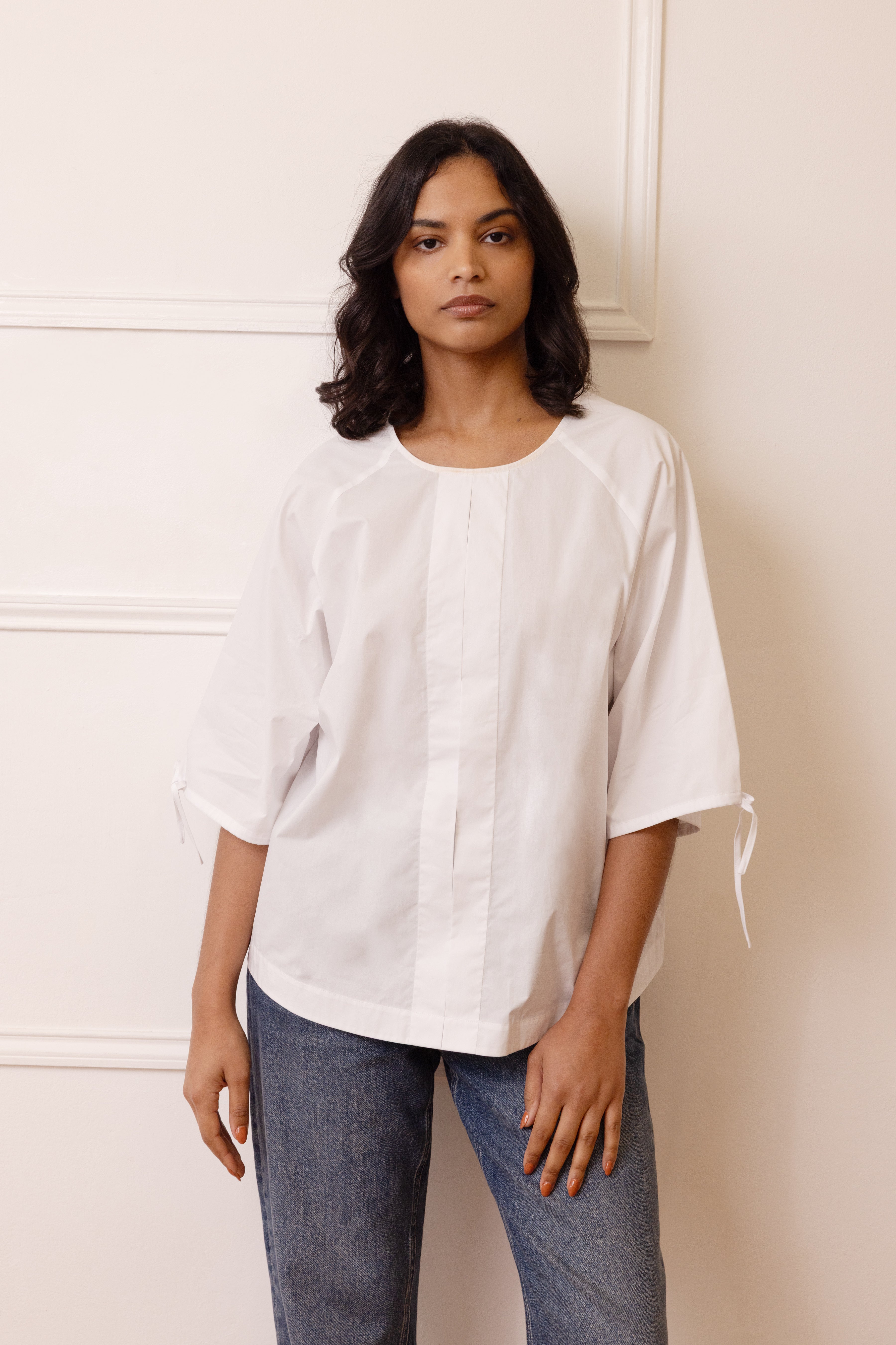 Woman wearing the Sierra Sleeve Tie Blouse in white. A relaxed sustainable cotton top with a central seam and adjustable tie details on the sleeves