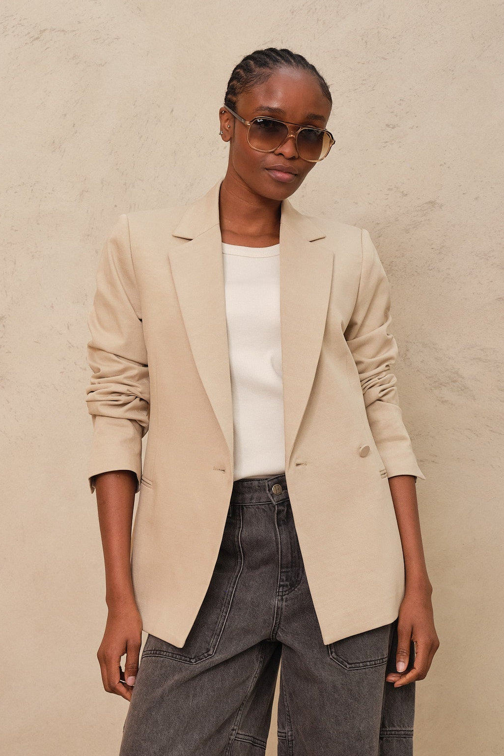Person wearing a beige blazer, white shirt, gray pants, and sandals against a beige background