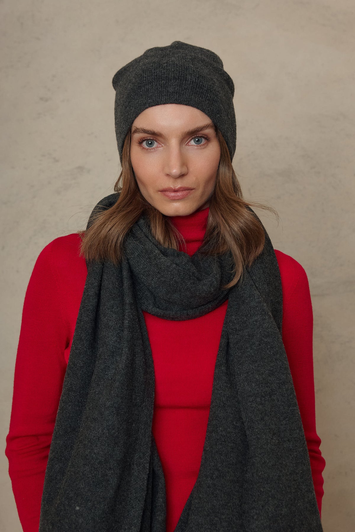 Model wearing the Carmel cashmere blend scarf in charcoal with the Zinnie beanie in charcoal and a red knit, front view