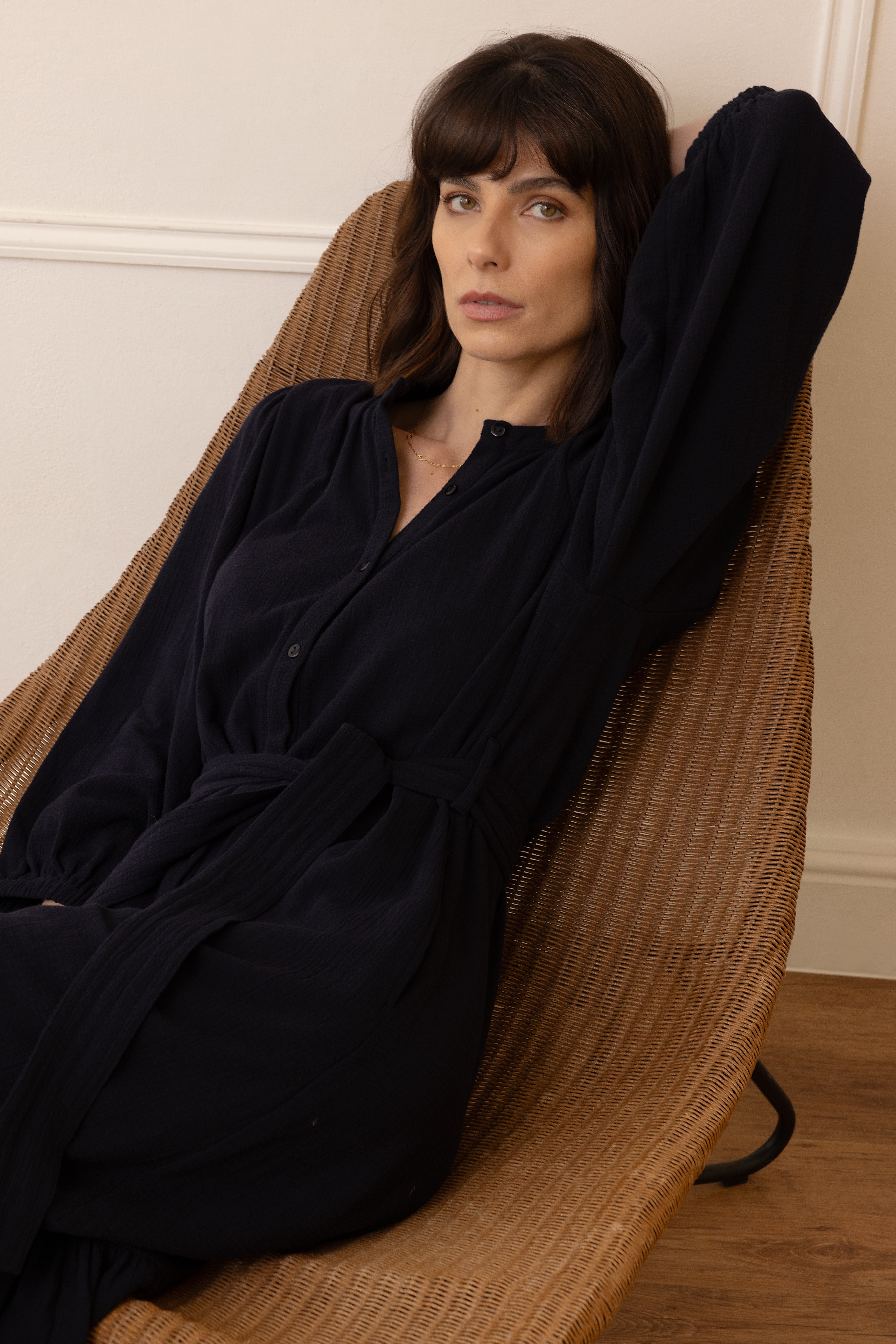Woman wearing a black dress standing in a room with a wooden floor and a wicker chair.