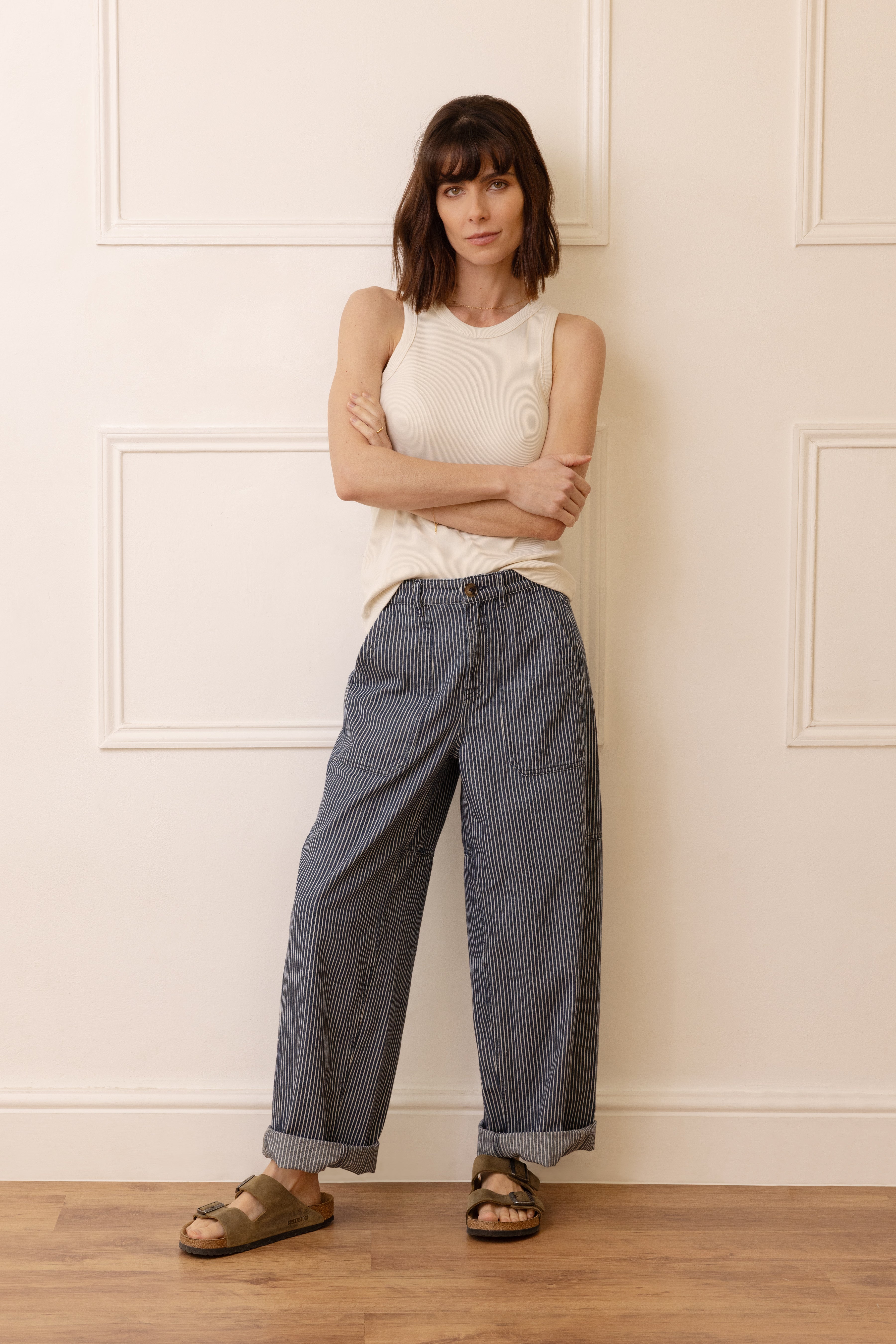 Woman wearing a white sleeveless top and blue striped pants standing against a white paneled wall.