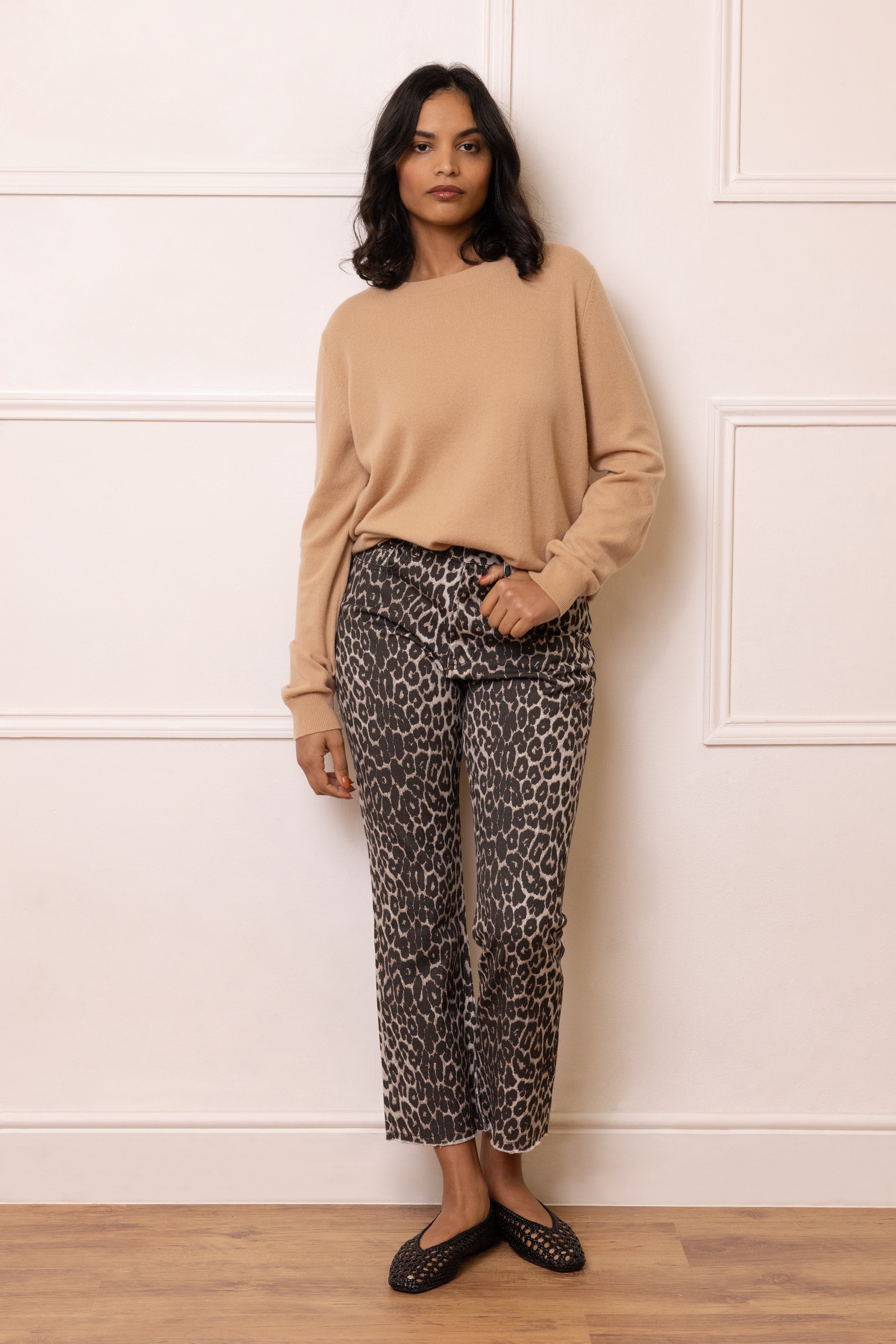 Woman wearing a beige sweater and leopard print pants against a white wall.