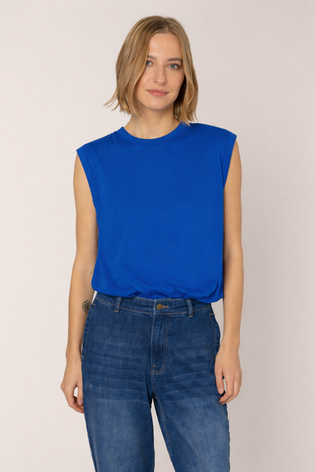 Woman wearing a blue sleeveless top and jeans against a plain background