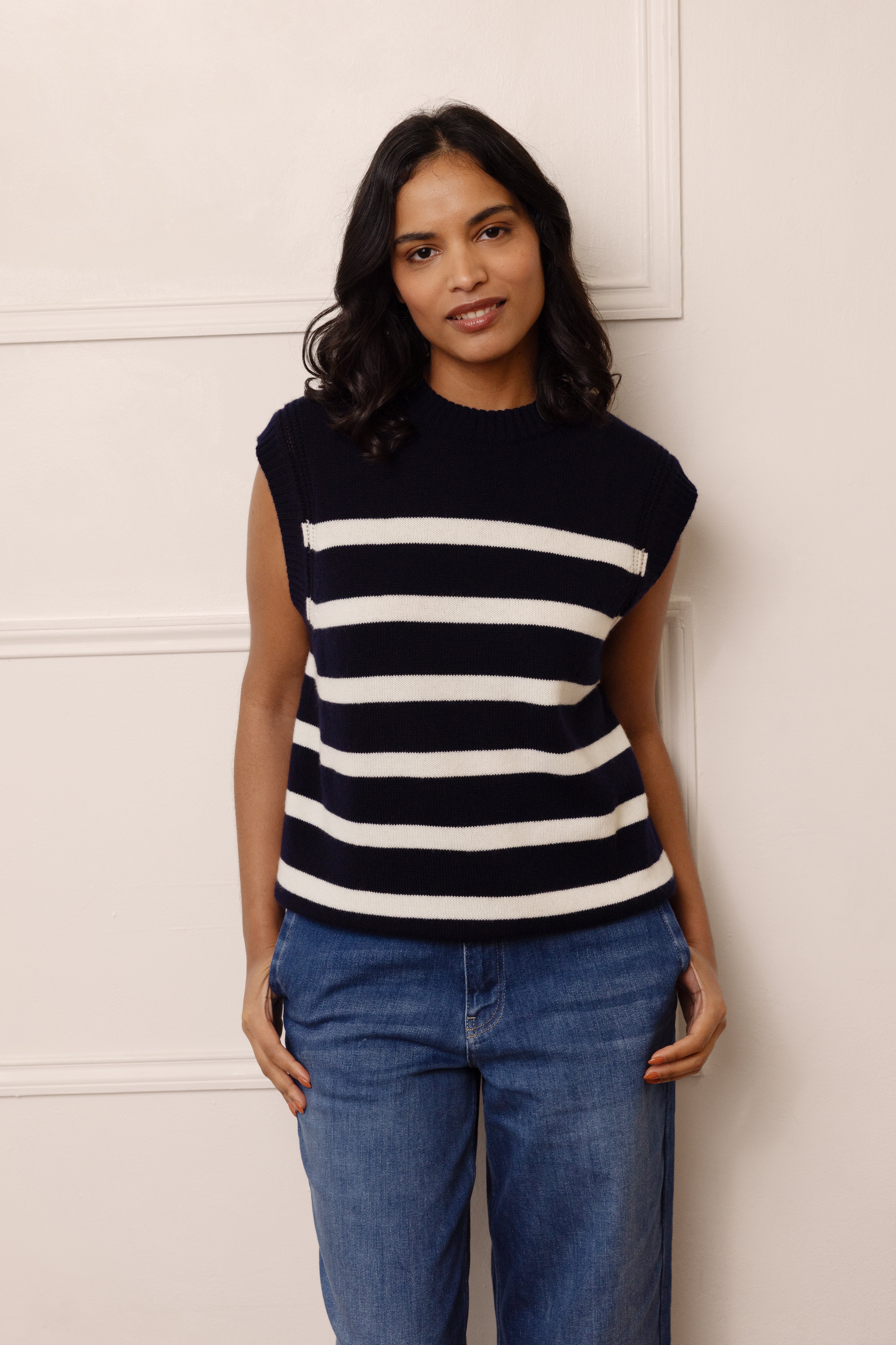Woman wearing a black and white striped sleeveless top and blue jeans against a light-colored wall.