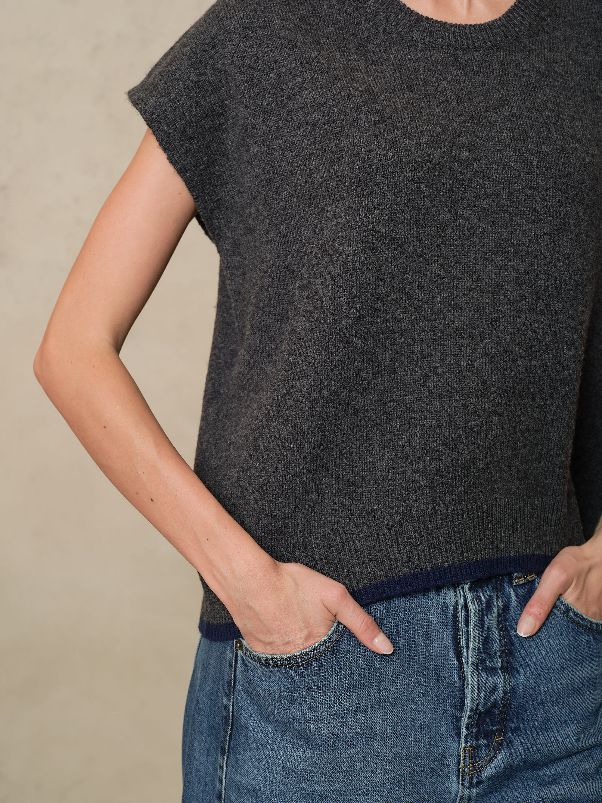Close view of the Ora charcoal knitted vest showing the ribbed hem worn over blue jeans