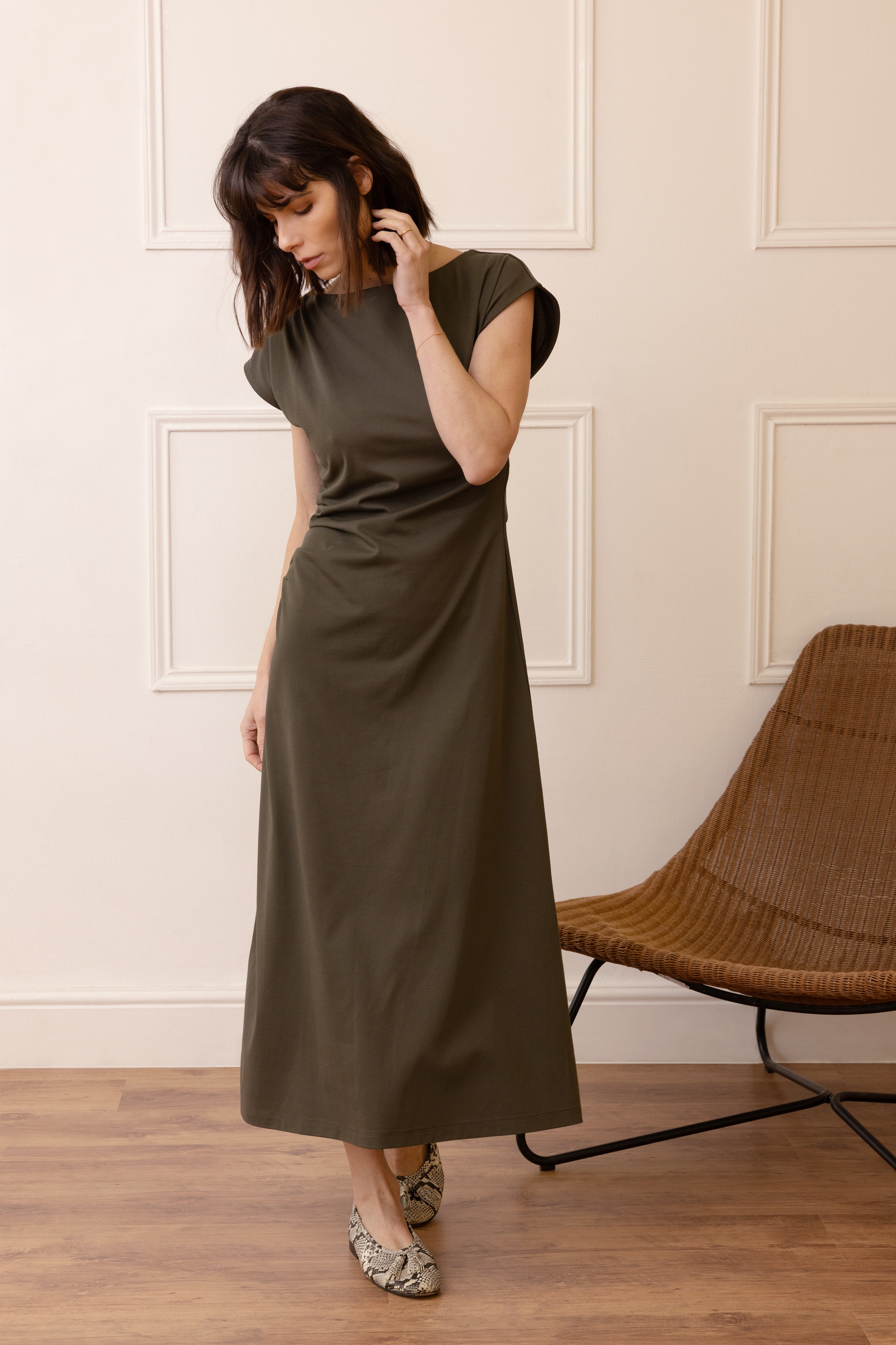 Woman wearing a long olive green dress standing in a room with wooden flooring and a wicker chair.