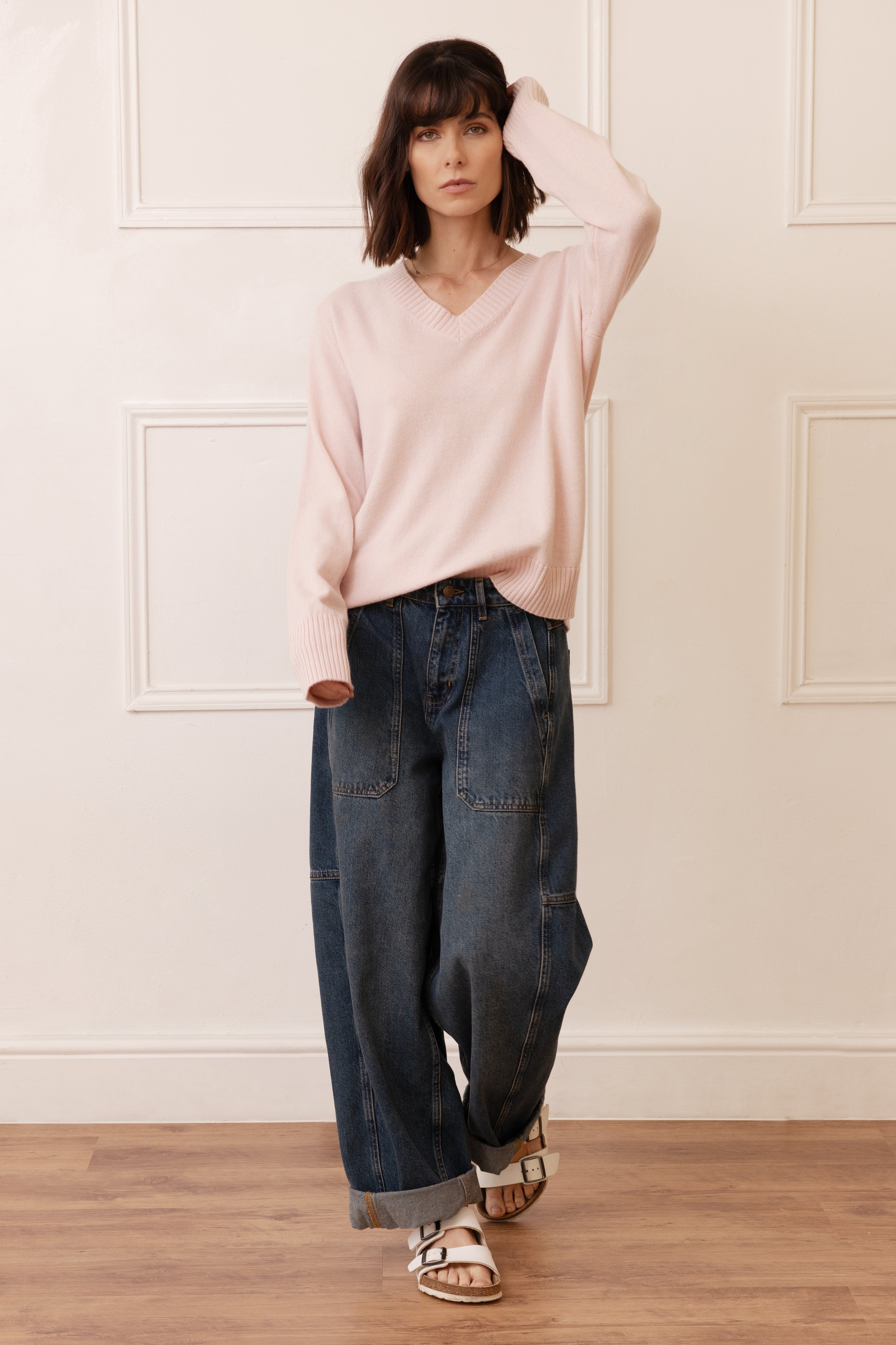 Woman wearing a light pink sweater and blue jeans standing against a white paneled wall.